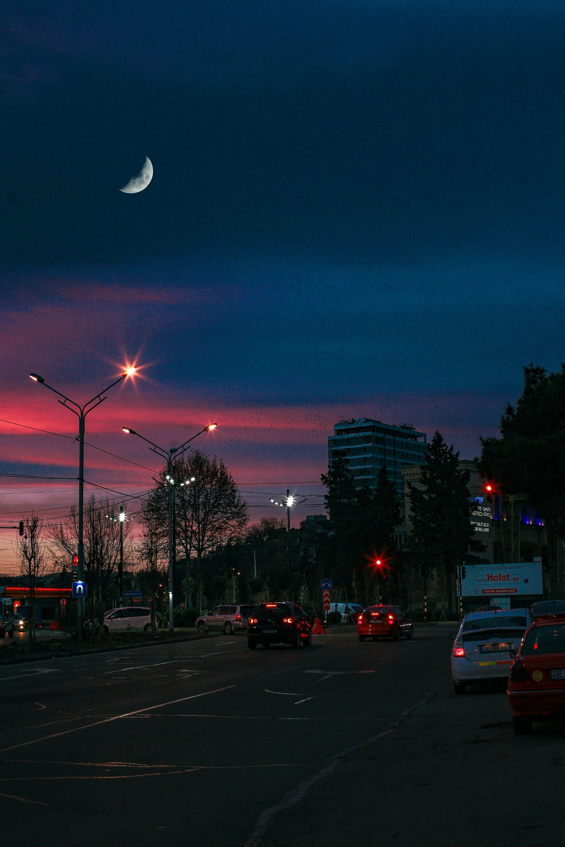 Roadway with vehicles near trees and buildings under colorful sky with moon in twilight in city district