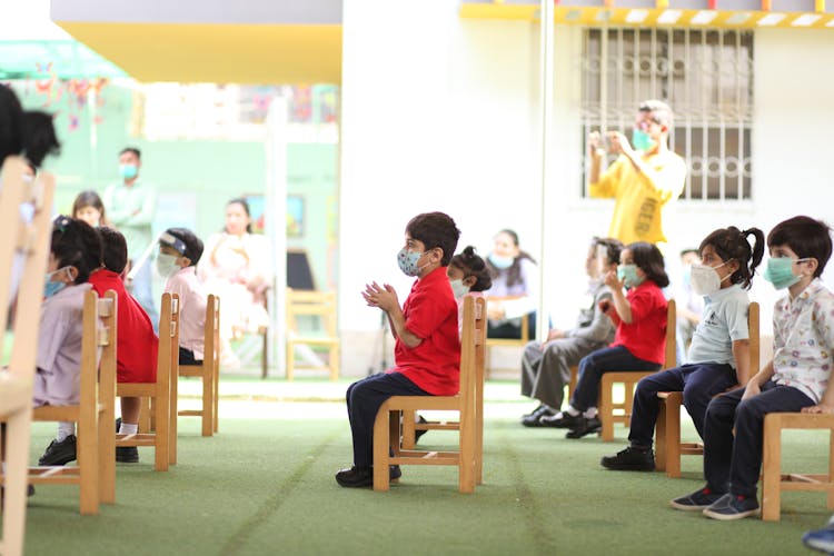 A Group Of Kids Sitting On Wooden Chairs While Wearing Face Mask