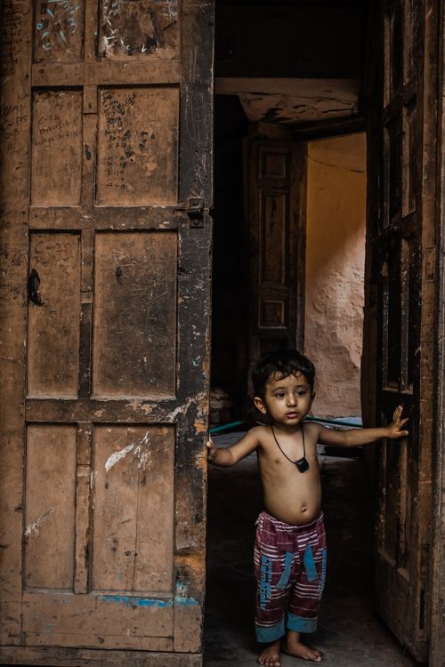 Free A young South Asian child stands innocently in a doorway, surrounded by rustic wooden textures. Stock Photo