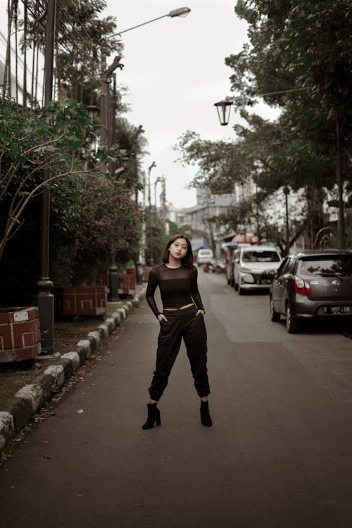 Full body of serious Asian female in stylish outfit standing confidently on roadway near parked cars