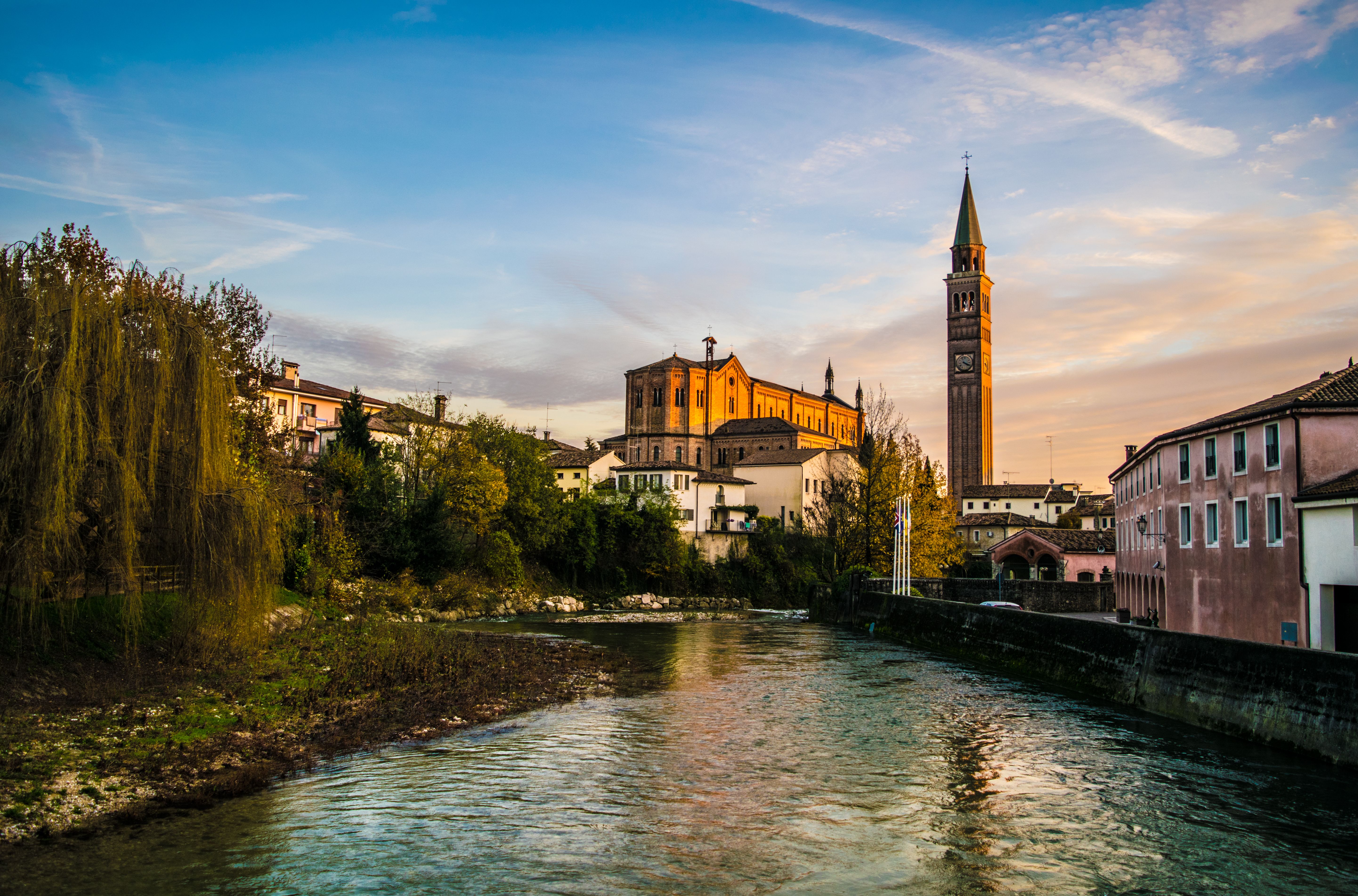 Picturesque view of Pieve di Soligo, Italy, featuring a church bell tower by a tranquil canal at sunset.
