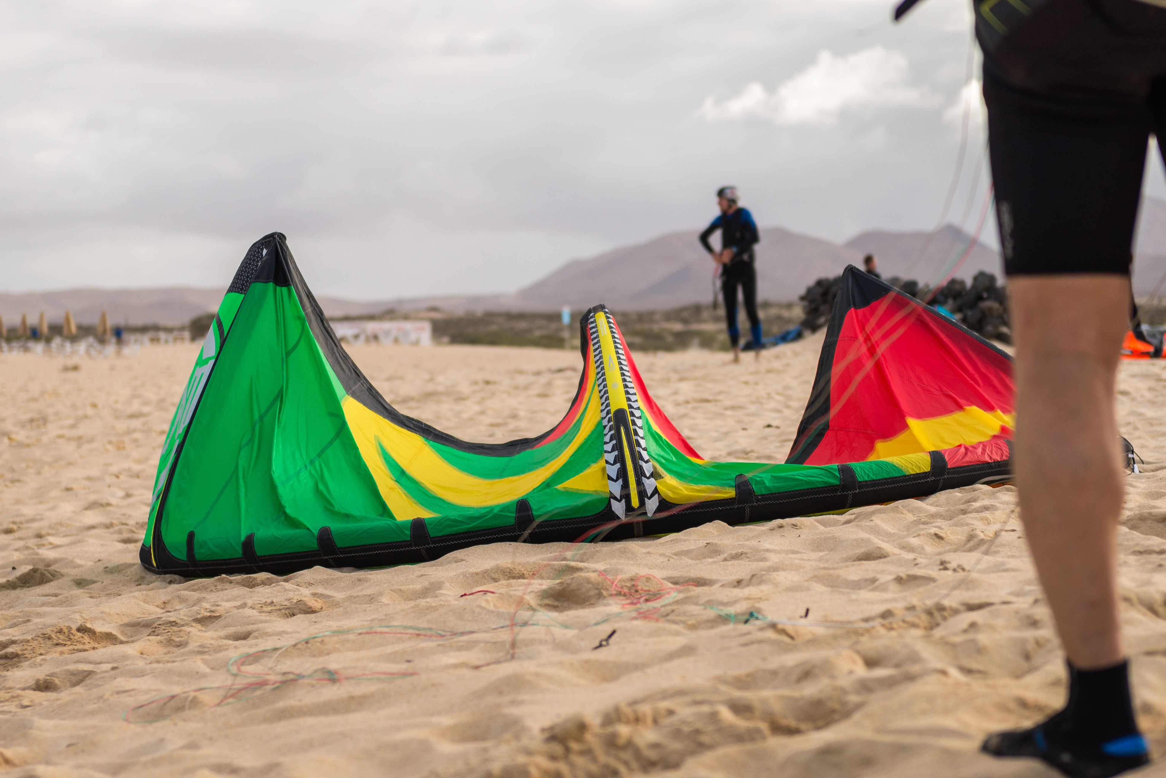Colorful kitesurfing equipment on a sandy beach with mountains in the background.