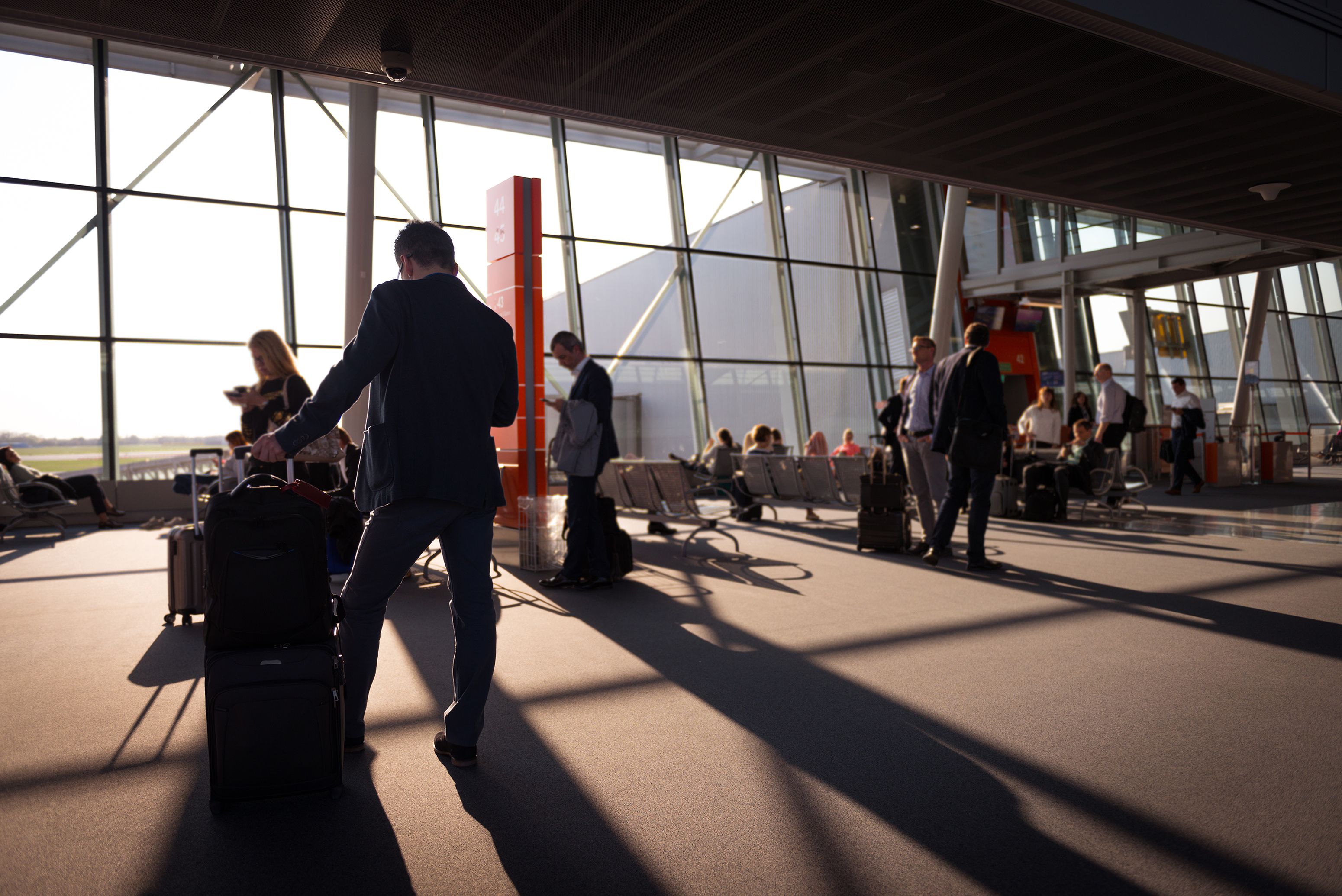 Travellers wait with luggage at an airport departure gate beside large windows overlooking the runway