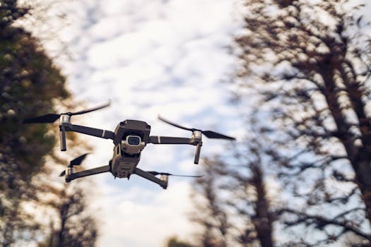 A drone flying close to the camera amid trees under a cloudy sky.