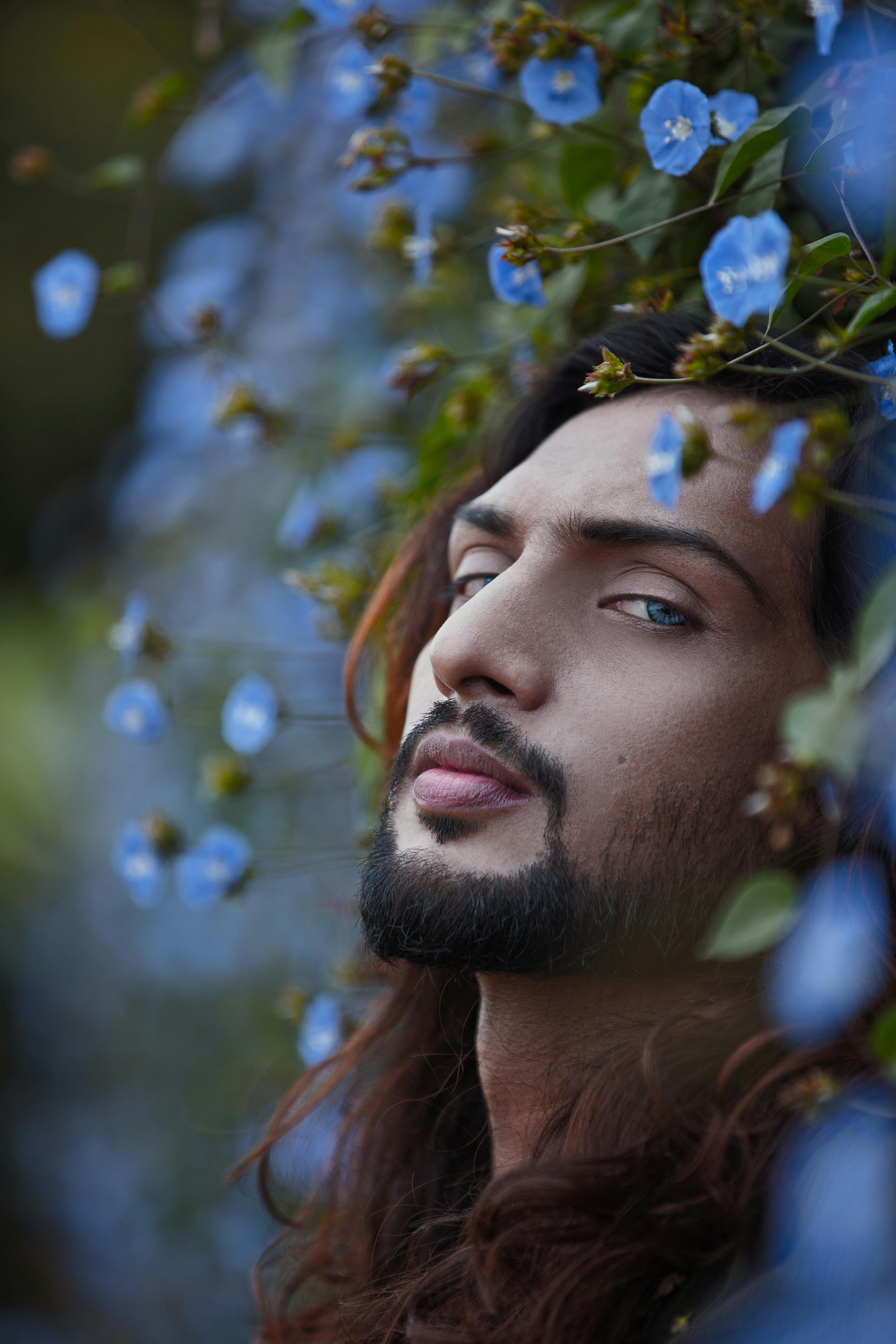 Close-up portrait of a bearded man surrounded by vibrant blue flowers, capturing a serene expression.