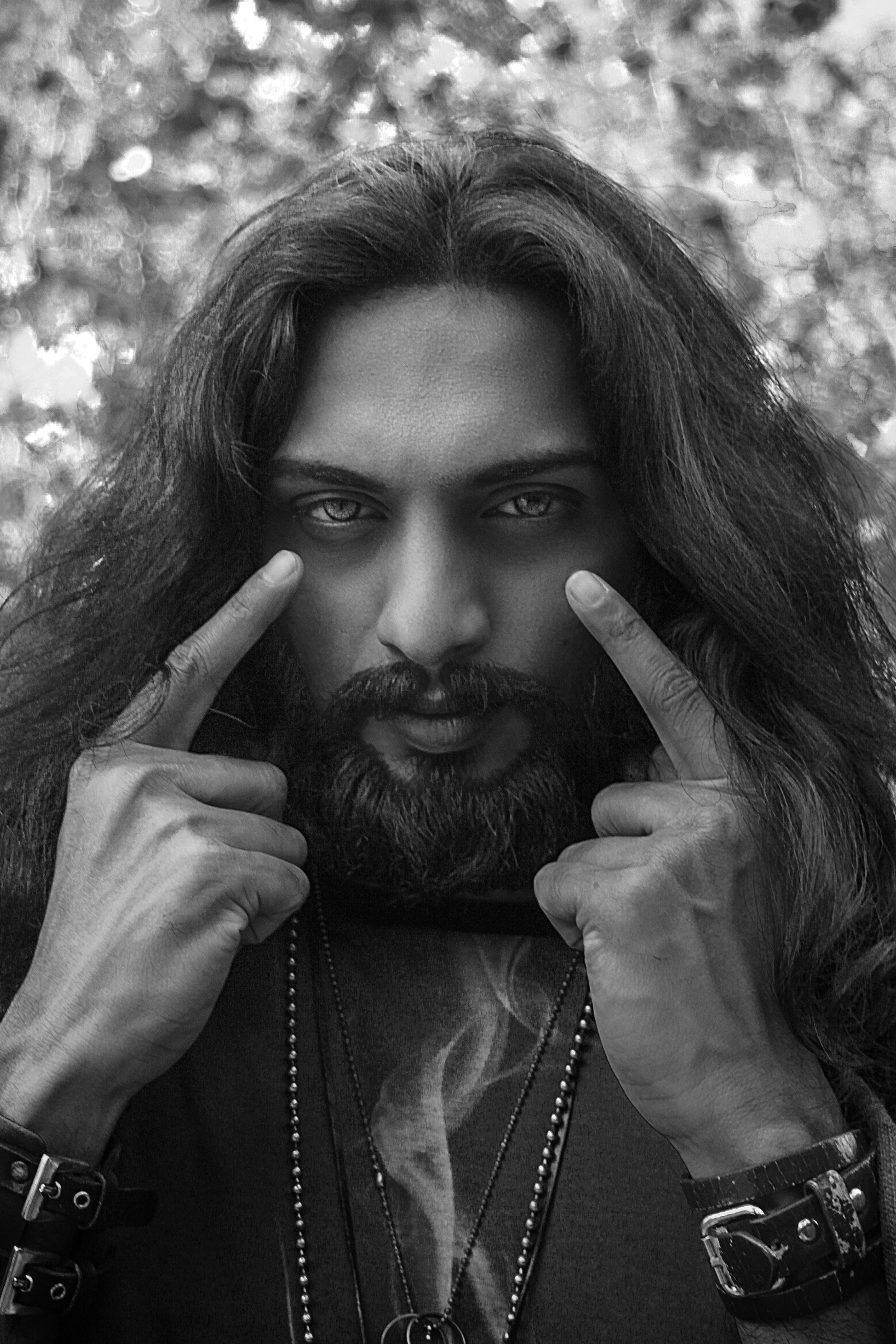 Close-up black and white portrait of a man with long hair and beard pointing to his eyes.