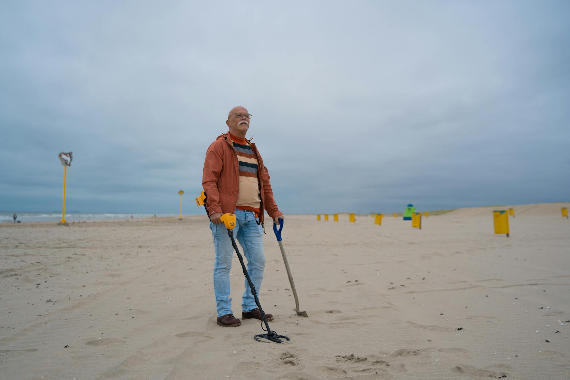 Elderly man with metal detector and shovel searching for treasures on a cloudy beach.