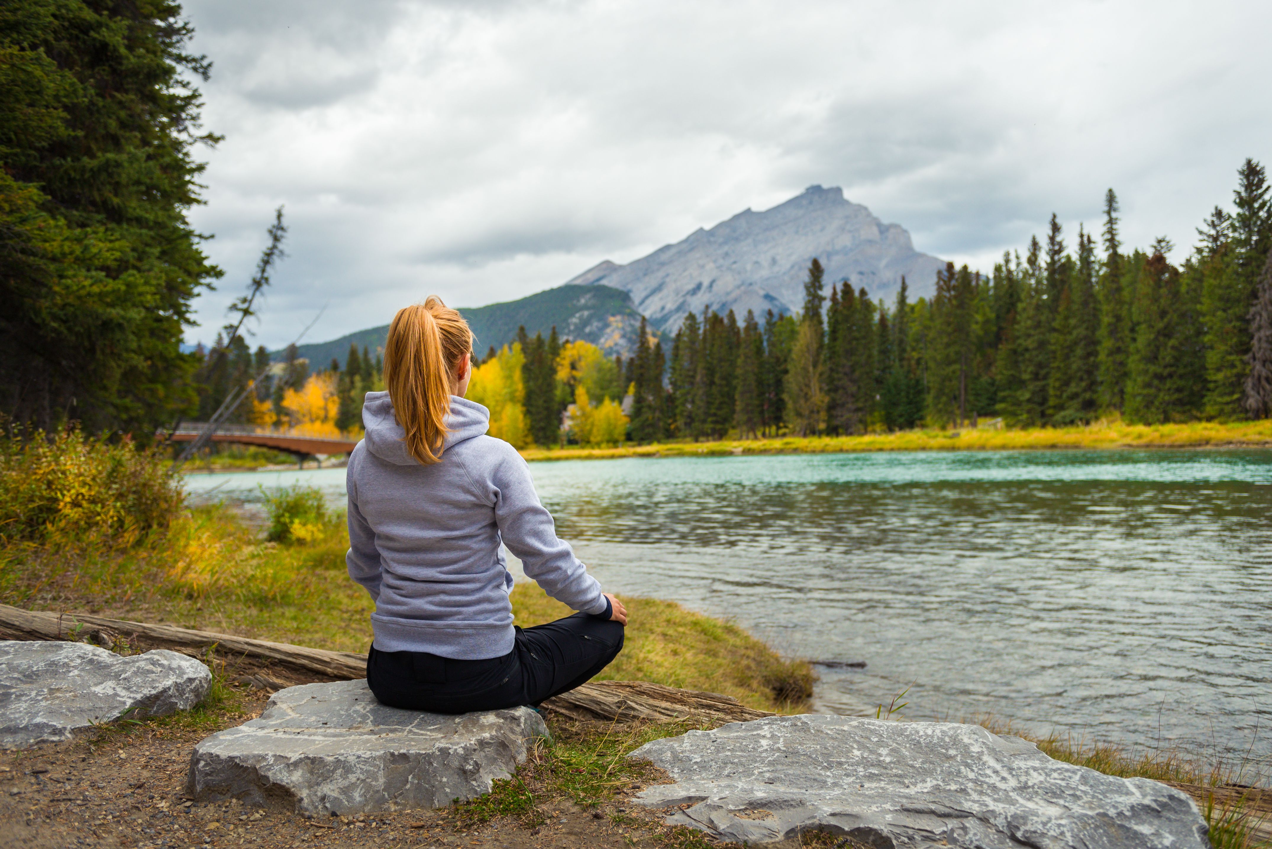 Woman meditating by a serene river with mountains in the background during autumn.