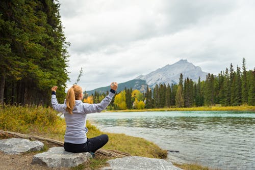 Woman in Grey Hoodie Sweater Sitting on Rock Near Body of Water