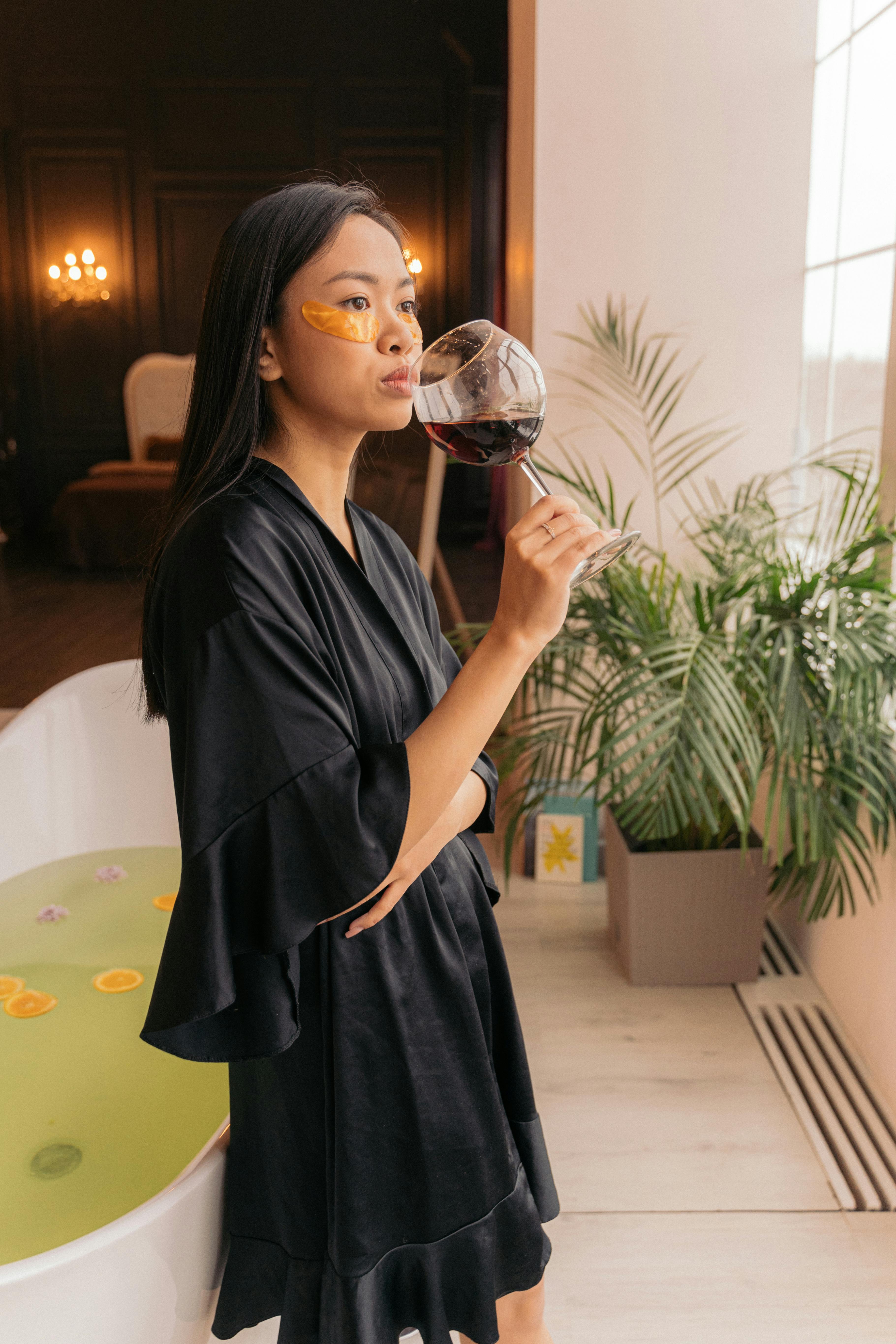 woman with under eye patches drinking wine next to bathtub
