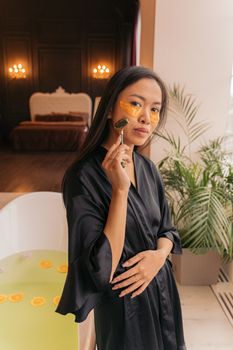 Woman in black robe uses facial roller and eye patches for skincare in a luxurious setting.