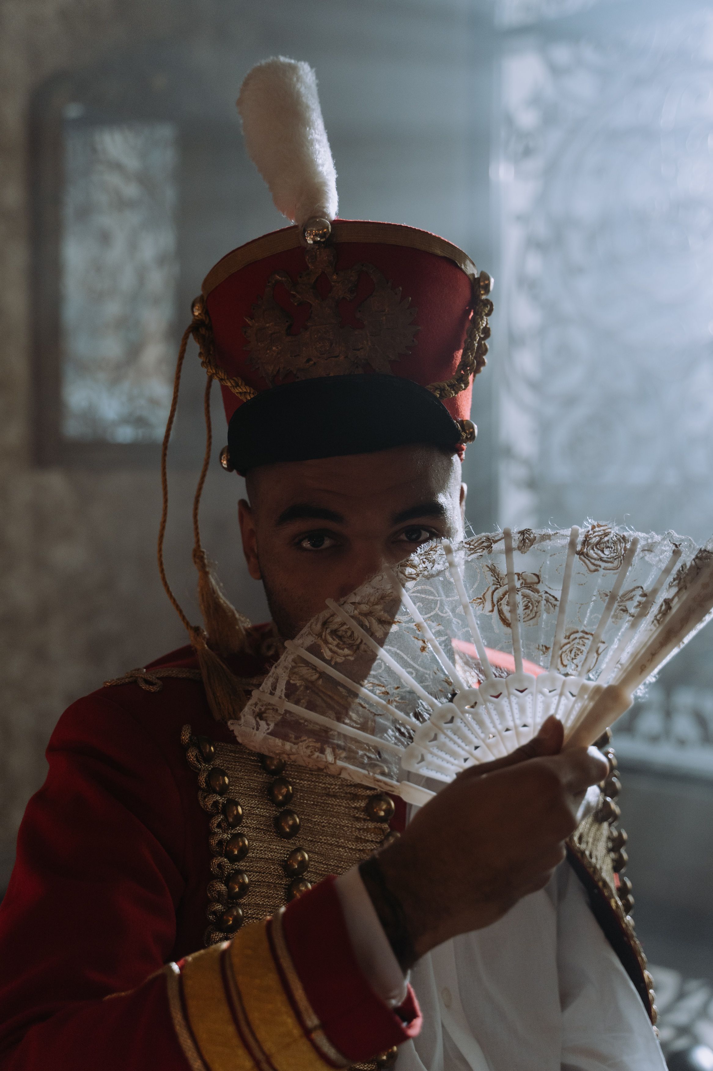 Man in historic costume with traditional red hat holding a decorative fan indoors.