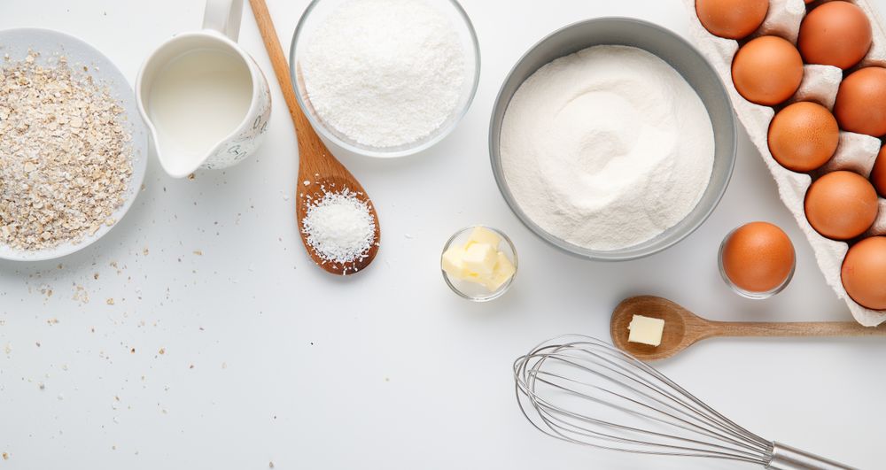 Baking ingredients with flour, milk, butter, oats, brown eggs, wooden spoons and a whisk on white