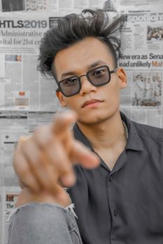 Stylish young Asian man in sunglasses pointing towards the camera against a newspaper backdrop.