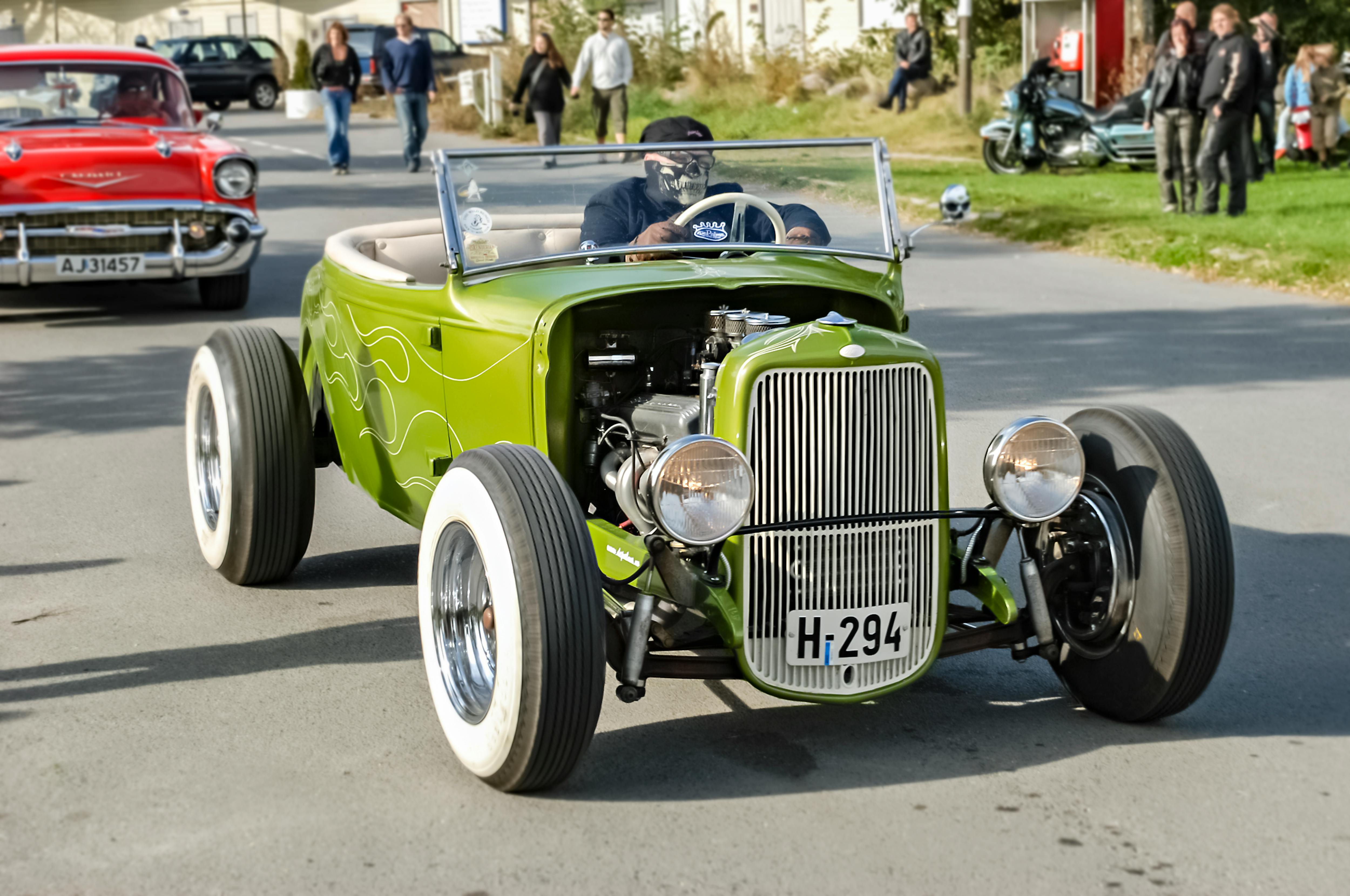 Vintage green hot rod car driving on a road with spectators in the background on a sunny day.