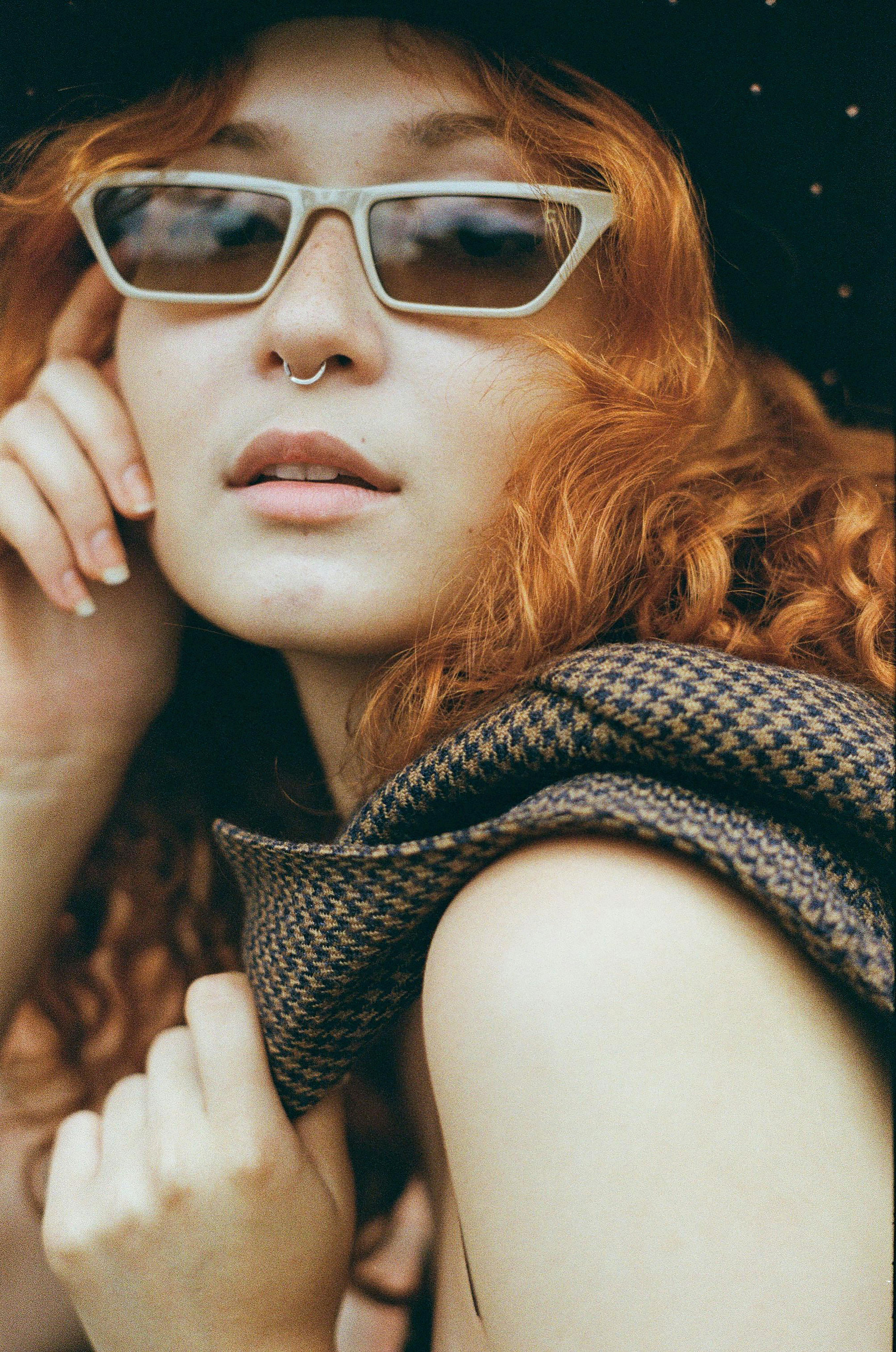 Trendy female with red hair and sunglasses with pierced nose wearing stylish black hat touching cheek while standing on street