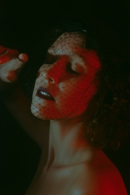 Sensitive female with closed eyes and bare shoulders wearing stylish black headdress with mesh standing in dark studio on black background