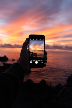 Hand holding smartphone capturing vibrant sunset over ocean in Tenerife.