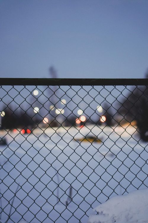 Free A snowy urban landscape viewed through a chain-link fence under evening lights. Stock Photo