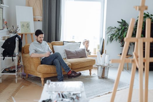 Casual man sitting on sofa using smartphone in a bright, modern art studio.