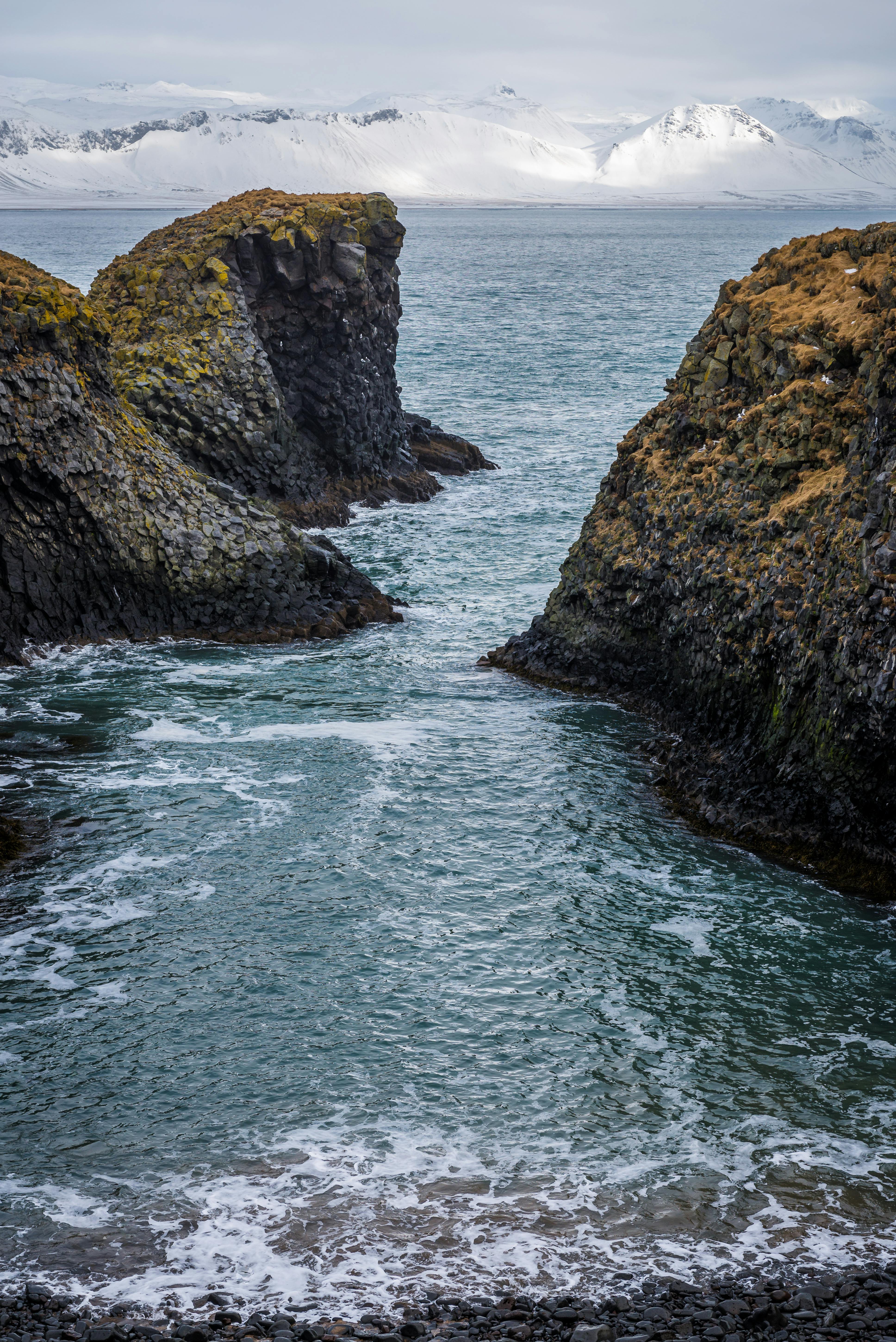 Dramatic coastal rock formations against snowy mountains and ocean waves create a stunning natural scene.