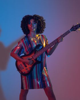 Stylish African American woman with curly hair playing bass guitar under colorful lights, exuding rock and roll vibes.