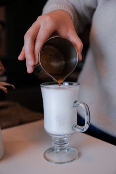 A close-up shot of a hand pouring espresso into a clear glass mug of milk latte.