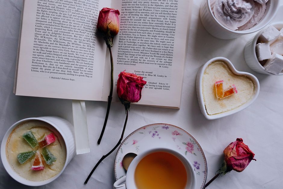 A flatlay featuring a book, cup of tea, colorful desserts, and dried roses.