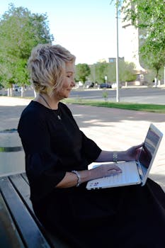 Side view of a woman with short hair typing on a laptop while sitting on an outdoor bench.