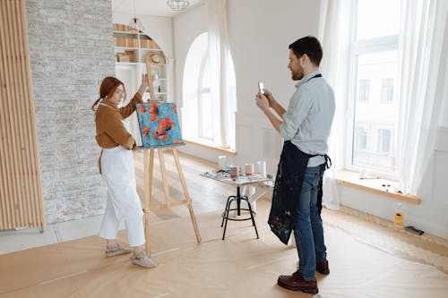 Man Taking Photo of the Woman Standing Beside the Painting 