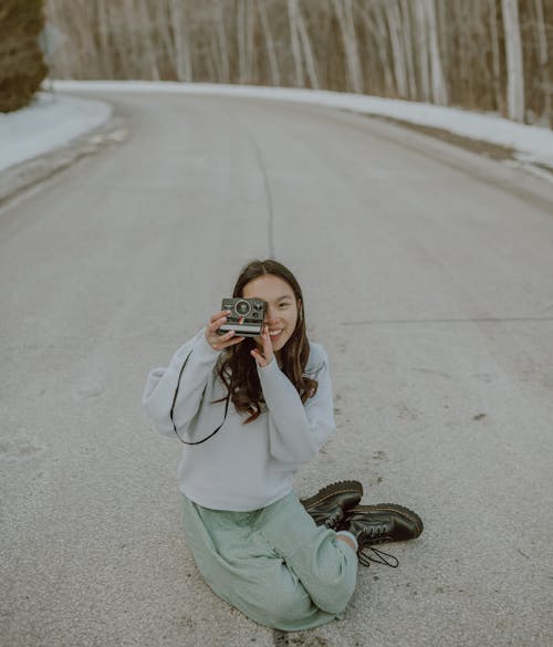 From above of happy young Asian female photographer in trendy outfit smiling and taking pictures on film camera sitting on asphalt road