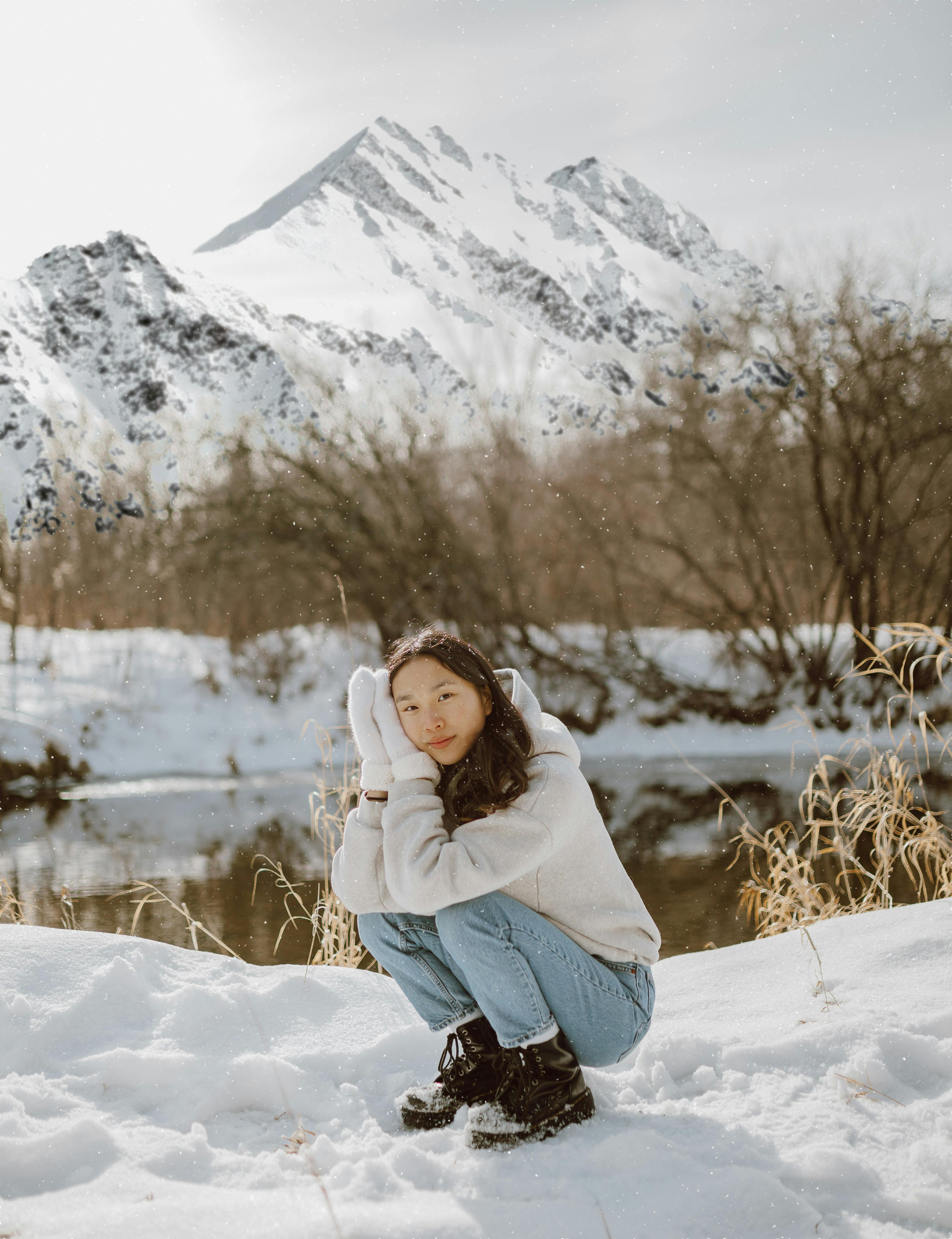Side view of relaxed young Asian female traveler with long dark hair in stylish warm clothes and mittens sitting on haunches on snowy ground near calm lake in mountainous valley