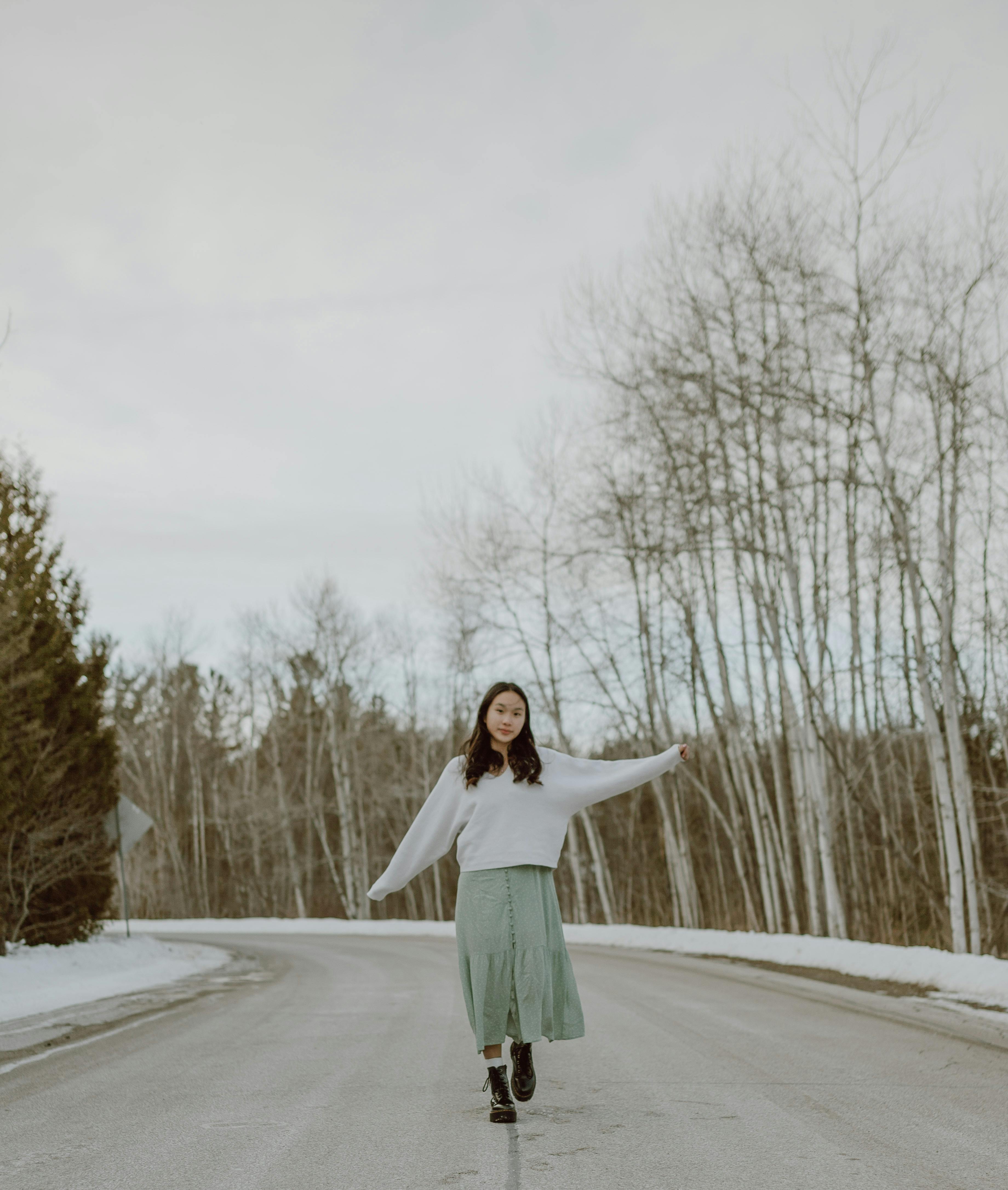 Full body of stylish young Asian female millennial with dark hair in trendy sweater and skirt walking along asphalt road with outstretched arms against cloudy sky