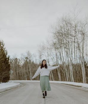 Full body of stylish young Asian female millennial with dark hair in trendy sweater and skirt walking along asphalt road with outstretched arms against cloudy sky