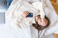 A man in a beige sweater holding a smartphone while lying on a bed indoors.