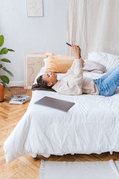 Woman lying on bed using smartphone in bright, cozy bedroom with plants and decor.