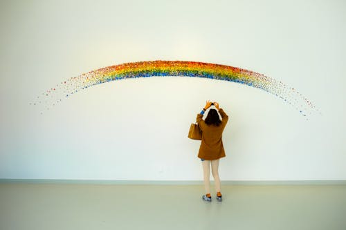 Женщина в коричневом топе и бежевых леггинсах фотографирует расписанную радугой стену