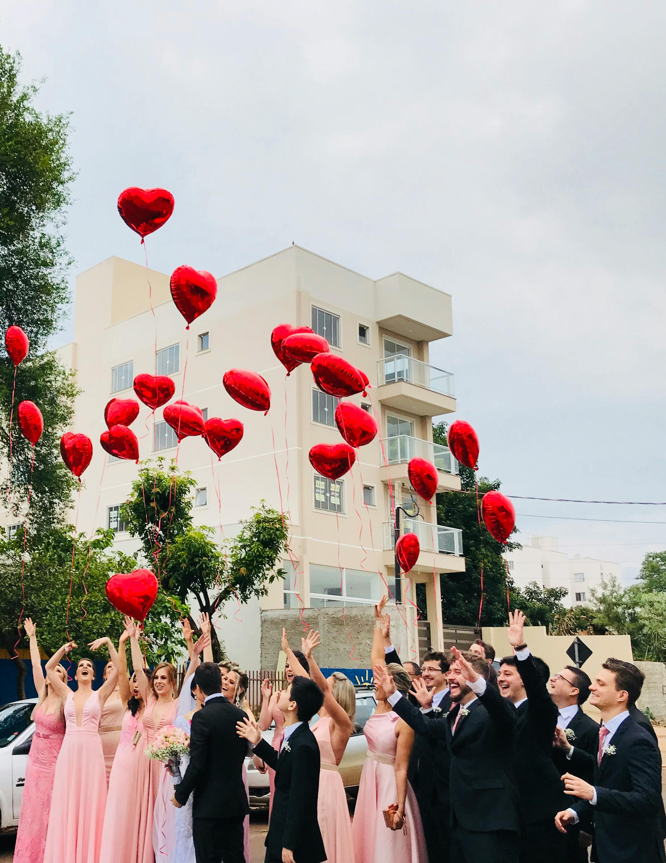 women wearing pink dresses and men wearing black suit jacket and pants raising hands with red heart balloons