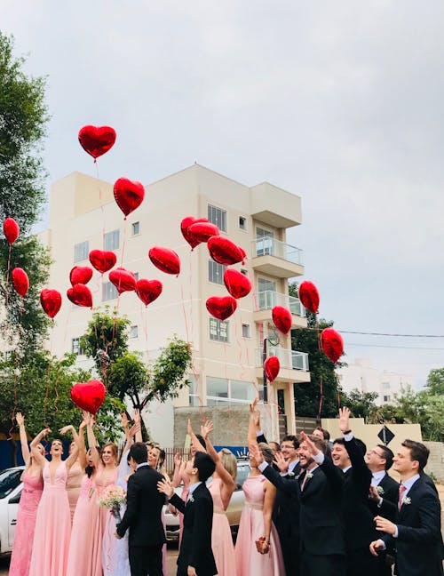Free Women Wearing Pink Dresses and Men Wearing Black Suit Jacket and Pants Raising Hands With Red Heart Balloons Stock Photo
