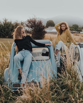 Two young women sitting on a vintage blue car amidst tall grass, exuding a relaxed and casual vibe.