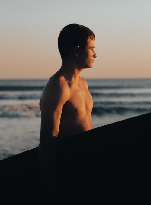 Free Shirtless young man with surfboard gazing at the ocean during sunset, embodying a serene coastal lifestyle. Stock Photo