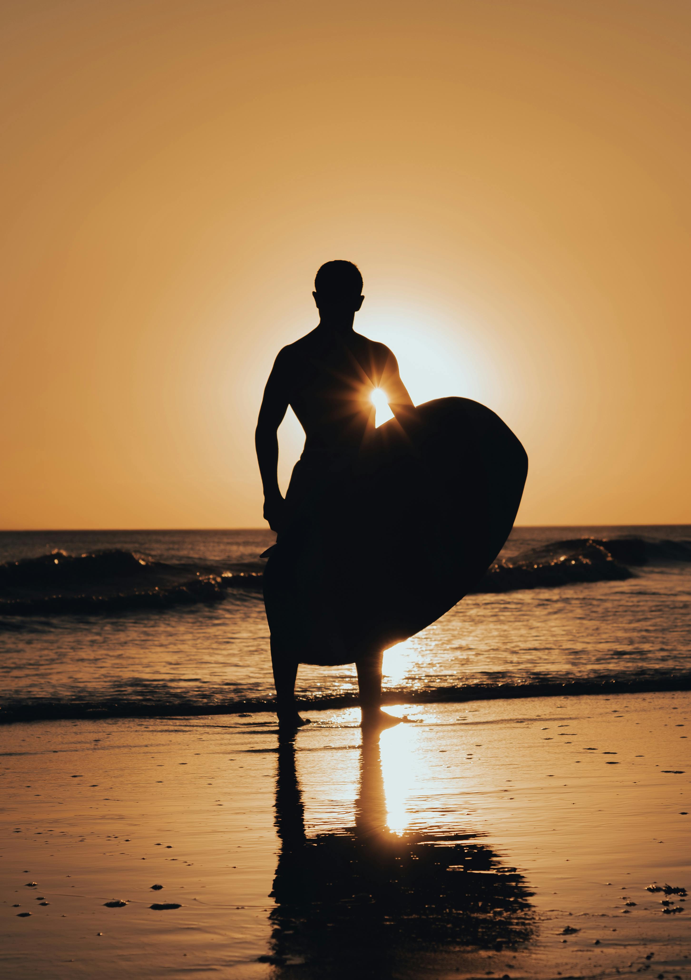 A silhouetted man with a paddle board stands against a stunning sunset backdrop on the ocean shore.