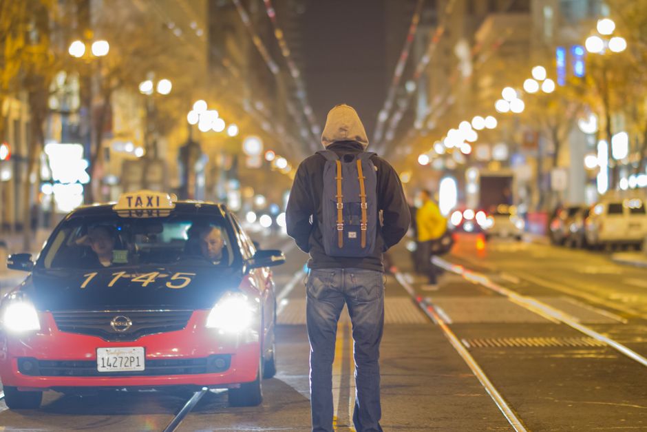 A person with a backpack stands in front of a taxi on a brightly lit city street at night.