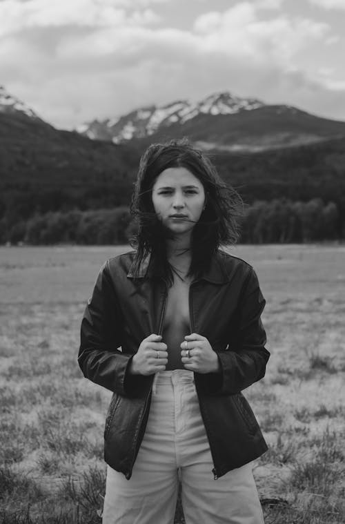Black and white of young thoughtful lady with dark hair in fashionable outfit standing in meadow near mountains and looking at camera on windy day