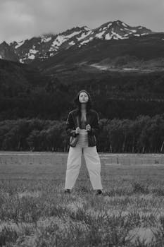 A young woman in a leather jacket stands confidently in a tranquil mountain meadow.