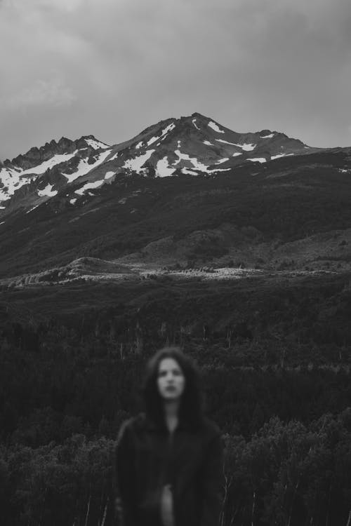 Black and white of unrecognizable young female tourist with long dark hair recreating in mountainous valley amidst lush trees under overcast sky