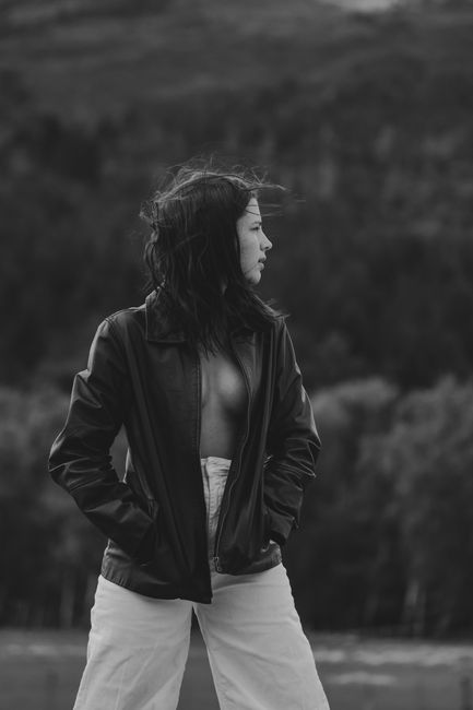 Black and white of dreamy young female tourist with long dark hair in trendy outfit admiring nature while standing near lush forest growing on mountain slope