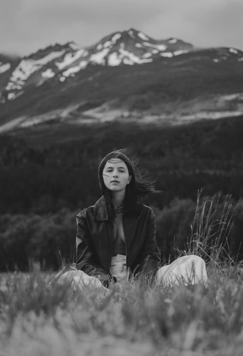 Free Black and white of romantic young female traveler with long hair in stylish clothes recreating on grassy meadow near mountain ridge against overcast sky on windy day Stock Photo