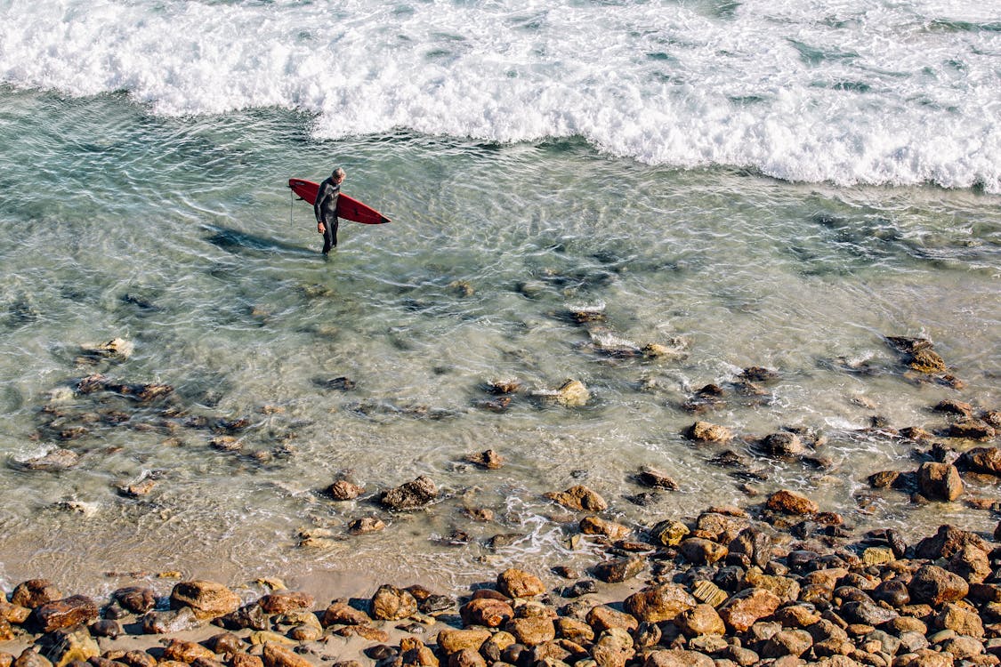 Bezpłatne Osoba Trzymająca Czerwoną Deskę Surfingową W Czystej Wodzie W Pobliżu Brązowego Kamienia W Ciągu Dnia Zdjęcie z galerii