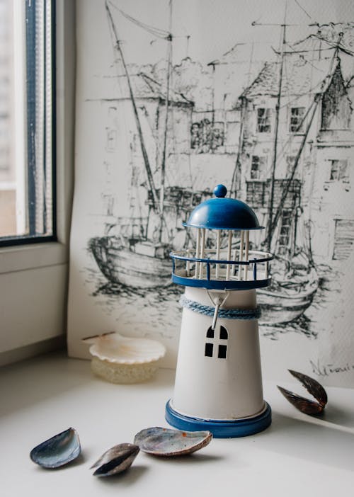 Composition of white and blue miniature lighthouse tower with creative pencil painting placed on table with scattered seashells near window